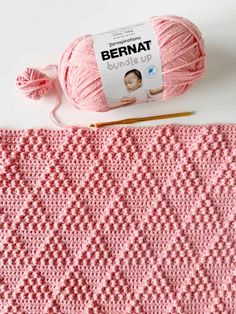 a pink crochet blanket next to a ball of yarn and a knitting needle