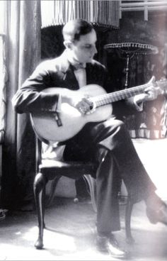 a man sitting in a chair holding a guitar and looking at the ground with his legs crossed