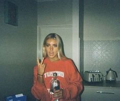 a woman in a red sweatshirt holding a bottle and making the peace sign with her hand