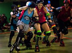 two women rollerblading down a track with other people in the background wearing helmets and knee pads