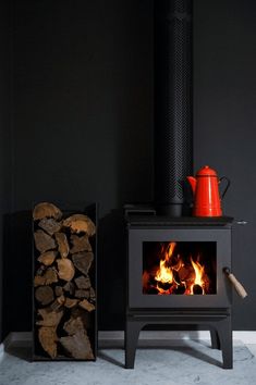 a wood burning stove next to a pile of firewood and a red teapot