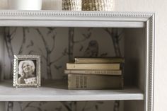 some books are sitting on a shelf in front of a wallpapered wall and a framed photo