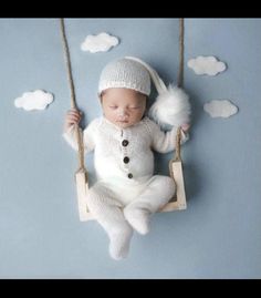 a baby in a white knitted outfit is sitting on a swing with clouds above him