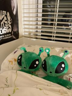 two green alien stuffed animals laying on top of a bed in front of a window