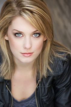 a woman with blonde hair and blue eyes wearing a black jacket is looking at the camera