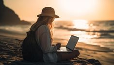 a woman sitting on the beach working on her laptop