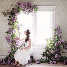 a woman sitting in front of a window surrounded by purple flowers