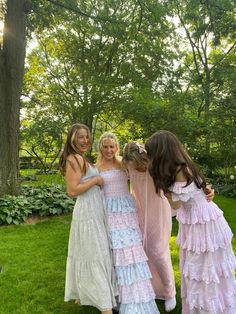 three girls in dresses standing next to each other on the grass with trees and bushes behind them