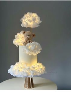 a teddy bear sitting on top of a white cake with fluffy clouds and lights around it