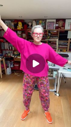 a woman in pink shirt and colorful pants standing on wooden floor next to bookshelves