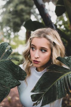 a woman with blonde hair and blue eyes is standing in the woods holding green leaves