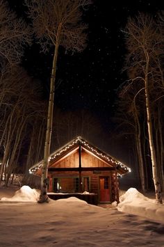 a small cabin is lit up with christmas lights in the snow and surrounded by trees