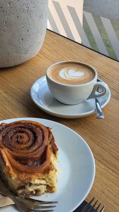 a piece of cinnamon roll on a plate next to a cup of cappuccino