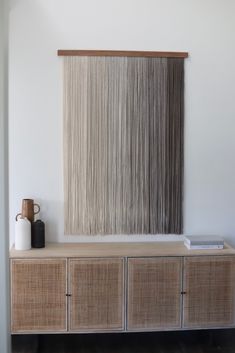 a wall hanging on the side of a white wall next to a wooden cabinet and vase