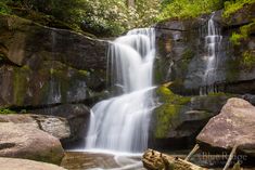 a waterfall in the middle of a forest with large rocks and trees around it's edge