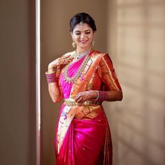 a woman in a pink and orange saree standing next to a wall with her hands on her chest