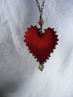 a red heart with pearls and beads hanging from a chain on a white cloth background