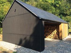 a garage with logs stacked in the front and side doors open on gravel area next to trees