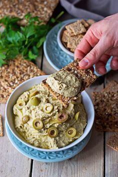 a hand dipping olives into a bowl of hummus with crackers on the side