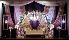 a couch sitting under a chandelier in front of purple drapes and flowers