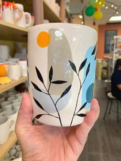 a hand holding up a coffee cup with leaves on it in front of shelves filled with plates and bowls