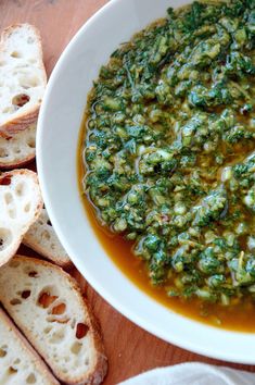 a white bowl filled with pesto next to slices of bread on a wooden table