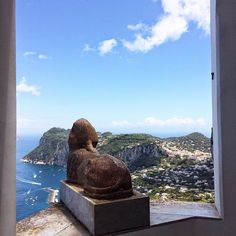 a statue sitting on top of a cement block next to the ocean and mountains in front of it