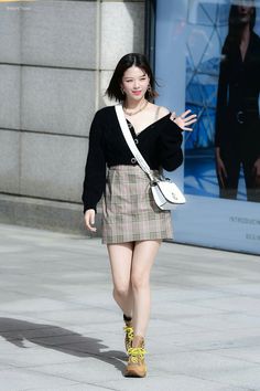 a woman walking down the street with her hand up to her chest, wearing a black sweater and plaid skirt