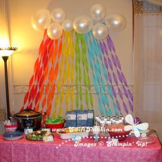 a table topped with lots of food next to a rainbow colored wall and balloons hanging from the ceiling