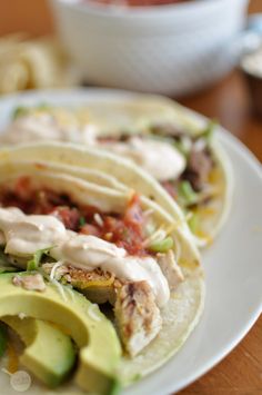 two tacos on a white plate with sauce and avocado in the background