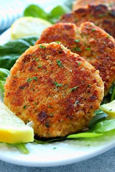 two crab cakes on a white plate with lemon wedges and spinach leaves next to it