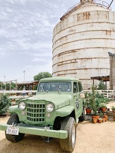 an old green truck is parked in front of a large silo with potted plants