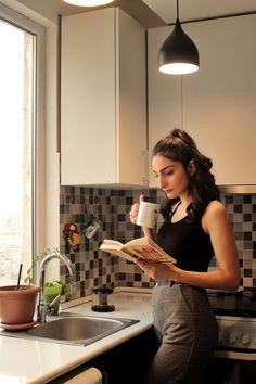 a woman standing in a kitchen reading a book and drinking from a cup while holding a coffee mug