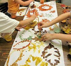 three children are painting leaves on paper at a table with oranges and yellows