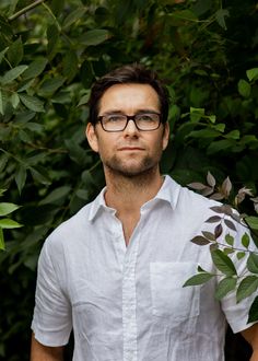 a man with glasses standing in front of some trees and bushes, looking at the camera