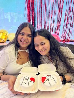 two young women smile as they hold plates with drawings on them, one is holding a heart