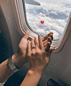 two hands reaching out an airplane window to touch the sky with a red heart on it