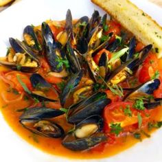 a white plate topped with mussels and tomato sauce next to a piece of bread