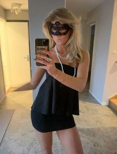 a woman in a black dress and mask taking a selfie with her cell phone