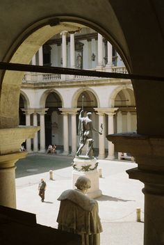 a statue in the middle of a courtyard with people walking around it and an archway leading to another building