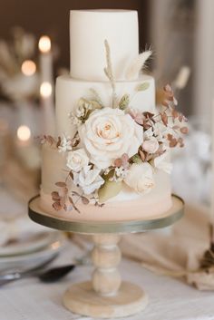 a wedding cake with white flowers and feathers on top is sitting on a table in front of candles