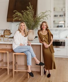 two women sitting on stools in a kitchen next to an island with potted plants