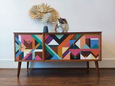 a cat sitting on top of a colorful cabinet with an art piece in the background