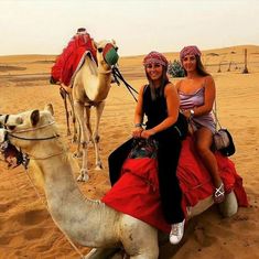 two women sitting on top of a camel in the middle of the desert with another camel behind them