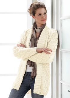 a woman leaning against a wall with her arms crossed wearing a sweater and scarf over her shoulders