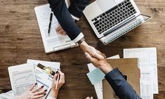 two people shaking hands over papers and laptops on a wooden table with other office supplies