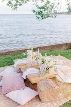 a picnic table set up on the grass with flowers, candles and plates in front of water