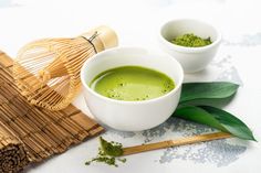 two white bowls filled with green liquid next to bamboo sticks and leaves on a table