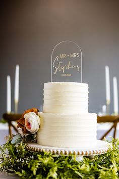 a white wedding cake with a mr and mrs sign on top, surrounded by greenery