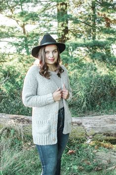 a woman standing in the woods wearing a black hat and cardigan sweater with long sleeves